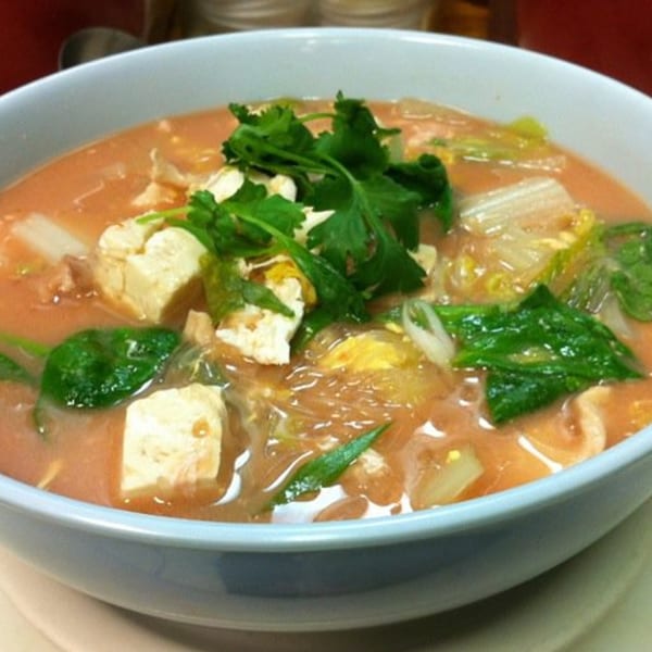 Close-up of a Thai soup with tofu, noodles, and green vegetables in a light blue bowl.